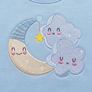 May include: A light blue fabric featuring an embroidered design of a smiling crescent moon wearing a blue and pink striped nightcap, accompanied by two smiling clouds and a small yellow star hanging from the moon by a thin pink line.