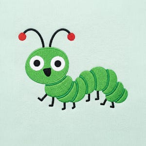 Può includere: Un ricamo raffigurante un bruco verde su un tessuto color menta chiaro. Il bruco presenta una testa rotonda con grandi occhi bianchi, pupille nere, una piccola bocca nera, due antenne nere con cerchietti rossi alle estremità e zampe nere.