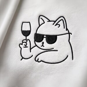Puede incluir: Una prenda de tela blanca con un diseño bordado en hilo negro que muestra una ilustración lineal de un gato con gafas de sol sosteniendo una copa de vino. El bordado está realizado con un hilo negro nítido y grueso sobre el fondo blanco.