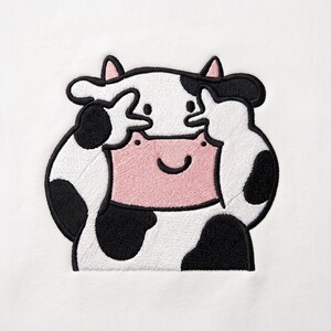 Peut inclure: Un écusson brodé sur un fond en tissu blanc représentant une vache de dessin animé avec des taches noires, des oreilles intérieures roses et un museau rose. La vache a ses sabots avant levés vers son visage, mimant un signe de paix.
