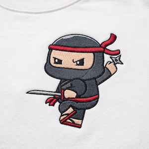 May include: A white t-shirt featuring an embroidered graphic of a cartoon ninja dressed in a black suit with red accents. The ninja is depicted in a dynamic pose, holding a silver sword in one hand and a four-pointed throwing star in the other.