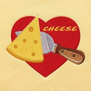 May include: A light yellow fabric featuring an embroidered red heart with the word CHEESE in yellow letters. A yellow triangular slice of Swiss cheese with holes sits on a silver metal cheese slicer with a brown handle over the heart.