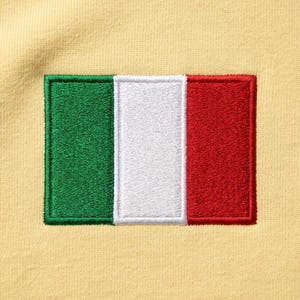 Pode incluir: Um emblema retangular bordado com a bandeira italiana, apresentando faixas verticais nas cores verde, branca e vermelha, costurado sobre um tecido amarelo claro. O bordado possui uma textura visível e uma borda definida ao redor do design.