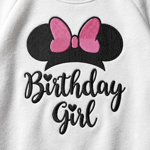 May include: A white fabric garment featuring an embroidered black silhouette of mouse ears with a bright pink bow. Below the ears, the text Birthday Girl is embroidered in black cursive script, with small heart icons above the i letters.