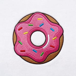 以下が含まれることがあります： 白い布地を背景にした、ドーナツ型の刺繍ワッペン。ドーナツは茶色のベースに、鮮やかなピンク色の波打つアイシングが施されており、赤、黄、緑、青、白の小さな長方形のカラースプレーが散りばめられています。