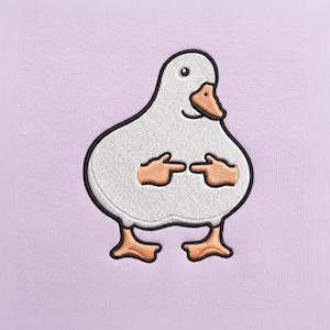 Op de afbeelding: Een lichtpaarse stoffen achtergrond met een geborduurde witte eend met een zwarte omlijning. De eend heeft een oranje snavel en oranje zwemvliezen en is afgebeeld met twee kleine oranje handjes die naar elkaar toe wijzen.