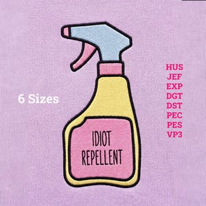 Può includere: Una toppa ricamata su tessuto viola chiaro raffigurante uno spruzzatore giallo con beccuccio azzurro e un'etichetta rosa. L'etichetta riporta il testo nero IDIOT REPELLENT. A sinistra 6 Sizes e a destra i formati file HUS JEF EXP DGT DST PEC PES VP3.
