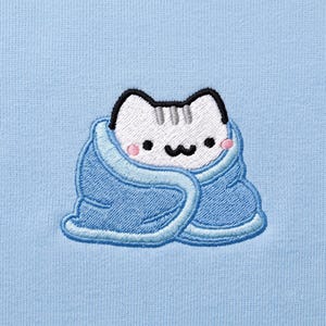 May include: A light blue fabric background featuring an embroidered graphic of a white cat with gray forehead stripes, black eyes, and a black mouth, wrapped snugly in a blue blanket with a lighter blue trim and small pink circular cheeks.