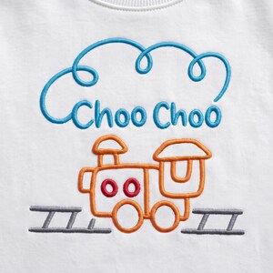May include: A white fabric garment featuring an embroidered design of an orange steam locomotive on grey tracks. Above the train, the text Choo Choo is stitched in blue, framed by a blue embroidered cloud-like outline above the lettering.