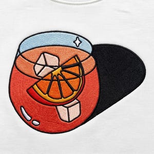 May include: An embroidered patch on a white fabric background featuring a round glass filled with a red liquid, ice cubes, and an orange slice garnish. The design includes a large black shadow extending to the right side of the glass.