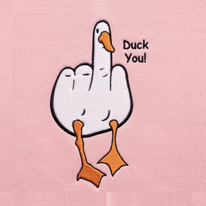 May include: A light pink fabric featuring an embroidered white goose with an orange beak and feet. The goose is shaped like a hand giving the middle finger. To the right of the goose, the text Duck You! is embroidered in black thread.