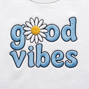Op de afbeelding: Een wit stoffen oppervlak met geborduurde tekst die good vibes leest in lichtblauw garen met een zwarte omlijning. De letter o in good is vervangen door een witte madeliefbloem met een geel hart en een zwarte omlijning.