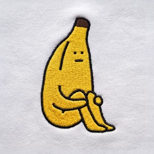 Pode incluir: Uma t-shirt branca com uma figura de banana amarela bordada na frente. A banana tem um caule castanho, contornos pretos e um rosto simples com dois pontos para os olhos e uma linha para a boca. A personagem está sentada com os joelhos.