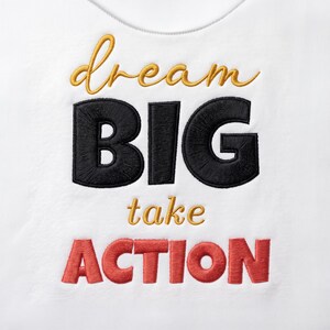 Può includere: Un indumento bianco con testo ricamato. La parola dream è in corsivo dorato, BIG in grandi lettere nere, take in corsivo dorato e ACTION in lettere rosse. Il ricamo è centrato sulla superficie del tessuto bianco.