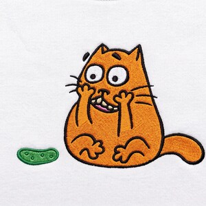May include: An embroidered patch on a white fabric background featuring a cartoon orange cat with wide eyes and a toothy grin, holding its paws to its face. To the left of the cat is a small, green, textured pickle shape with dark green dots.