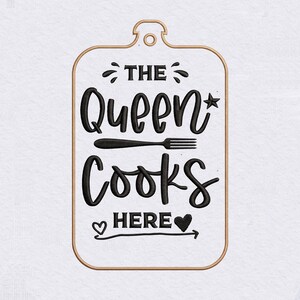 Może przedstawiać: Prostokątna biała naszywka z zaokrąglonymi rogami i miedzianą obwódką, zawierająca czarny maszynowy haft z napisem THE QUEEN COOKS HERE, uzupełniony ikoną widelca, małą gwiazdką oraz dwoma symbolami serca.