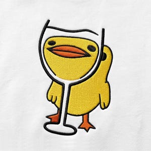 May include: An embroidered patch on a white fabric background featuring a bright yellow duckling with orange feet and a beak. The duckling is positioned behind a black-outlined white wine glass graphic that covers its face and upper body.