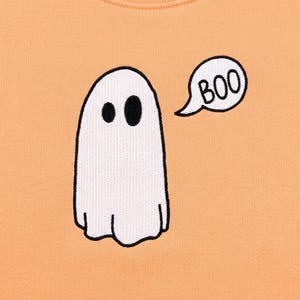 May include: A light orange fabric featuring an embroidered white ghost with black eyes and a black outline. To the right of the ghost is a white speech bubble with a black outline containing the black embroidered text that reads BOO.