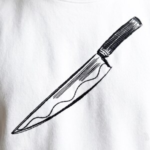 Peut inclure: Un vêtement en tissu blanc orné d'une broderie noire représentant un couteau de cuisine. La broderie montre une lame tranchante avec un détail de ligne ondulée et un manche texturé, positionnée en diagonale au centre de la surface textile.