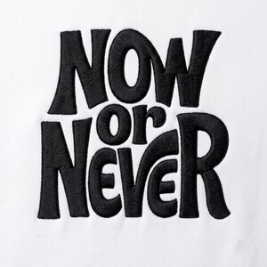 May include: A white fabric surface featuring black embroidered text that reads NOW OR NEVER in a bold, stylised, sans-serif font. The embroidery has a visible thread texture and creates a high-contrast design against the plain white background.