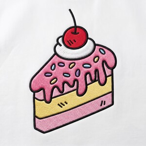 Peut inclure: Un écusson brodé représentant une part de gâteau sur fond blanc. Le gâteau présente une couche de génoise jaune, une couche rose et un glaçage rose avec des vermicelles colorés. Une touche de crème blanche et une cerise rouge ornent le sommet.