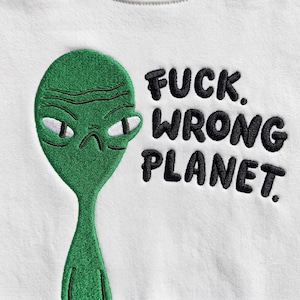 Può includere: Un capo in tessuto bianco con il ricamo di un alieno verde dall'espressione accigliata e gli occhi bianchi. A destra dell'alieno, il testo FUCK. WRONG PLANET. è ricamato in lettere maiuscole nere, spesse e arrotondate.