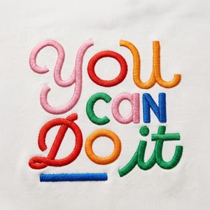 Könnte beinhalten: Eine weiße Stoffoberfläche mit dem gestickten Text "YOU CAN DO IT" in einer verspielten, mehrfarbigen Schriftart. Die Buchstaben sind mit rosa, roten, orangen, grünen und blauen Fäden gestickt, was ein lebendiges und strukturiertes Aussehen erzeugt.