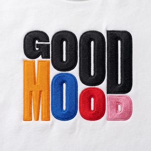 Puede incluir: Una superficie de tela blanca con el texto bordado GOOD MOOD. La palabra GOOD está cosida con hilo negro. La palabra MOOD está cosida con hilo multicolor, con la M en naranja, la O en azul, la O en rojo y la D en rosa claro.