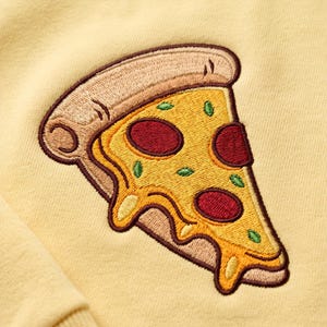 May include: A light yellow sweatshirt featuring a detailed embroidered pizza slice. The pizza has a tan crust, melted yellow cheese, dark red pepperoni circles, and small green basil leaves, all outlined in a dark brown thread.