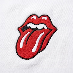 Può includere: Una superficie in tessuto bianco con una toppa ricamata centrale raffigurante una bocca rossa, nera e bianca con la lingua di fuori. Il design è composto da labbra rosse spesse, denti bianchi e una lingua bianca, il tutto contornato da un filo nero.