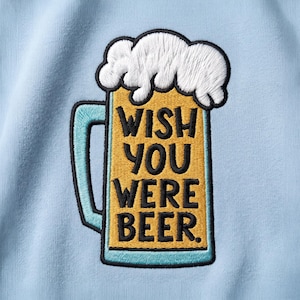 Peut inclure: Un sweat-shirt bleu clair avec un écusson brodé représentant une chope de bière avec de la mousse blanche. La chope est jaune avec une anse et une base bleu clair. Le texte noir indique, WISH YOU WERE BEER. La broderie a un contour noir épais.