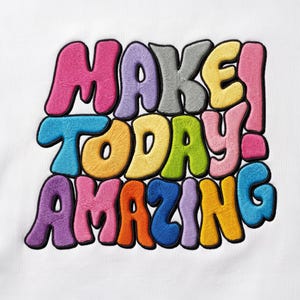 May include: A white fabric surface featuring a multi-colored embroidered design with the text "MAKE! TODAY AMAZING". The letters are rendered in a puffy, rounded style with black outlines, using vibrant shades of pink, purple, gray, yellow, blue, green, and orange thread.