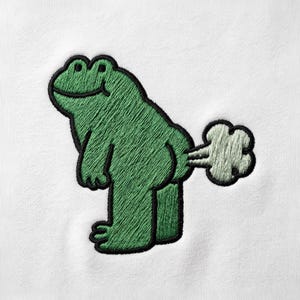 Puede incluir: Un parche bordado sobre una superficie de tela blanca que muestra una rana verde de perfil. La rana tiene un contorno negro y se representa con una forma de nube verde claro que sale de su parte trasera, sugiriendo una flatulencia.
