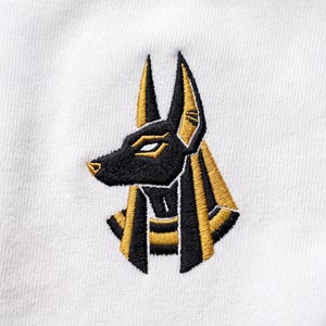 May include: A white fabric garment featuring a detailed embroidery of the Egyptian deity Anubis. The design depicts the jackal-headed figure in profile, rendered in black and metallic gold thread, showcasing pointed ears and a traditional headdress.
