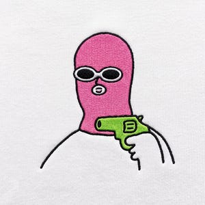 May include: An embroidered graphic on a white fabric surface featuring a pink balaclava-clad figure wearing white-rimmed oval sunglasses. The figure is holding a bright lime green revolver, outlined in black thread, pointing toward the right side.