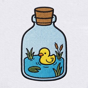May include: An embroidered patch featuring a glass bottle shape with a cork stopper. Inside the bottle is a blue pond with a yellow rubber duck, a green lily pad, green grass blades, and two brown cattails, all outlined in black thread.