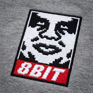 May include: A grey fabric garment featuring a rectangular embroidered patch. The patch displays a black and white pixelated face graphic above a red rectangular section containing the white embroidered text 8BIT in a blocky font style.