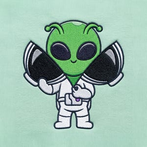 May include: An embroidered patch on a light mint green fabric featuring a green alien with large black eyes wearing a white space suit. The alien is holding two large black and white axes, one in each hand, and has a small utility belt around its waist.