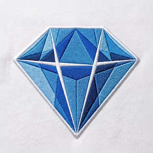 May include: An embroidered patch featuring a diamond shape in various shades of blue, including light, medium, and dark tones. The design is outlined in white thread and set against a plain white fabric background, showcasing geometric facets.