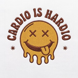 May include: A white fabric surface featuring an embroidered design of a yellow melting smiley face with brown outlines, X-shaped eyes, and a tongue sticking out. The text CARDIO IS HARDIO is arched above the face in brown capital letters.