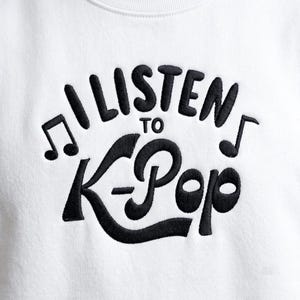 May include: A white sweatshirt featuring black embroidered text that reads I LISTEN TO K-Pop. The design includes two musical note symbols, one on the left and one on the right, flanking the text. The embroidery has a raised, textured appearance.