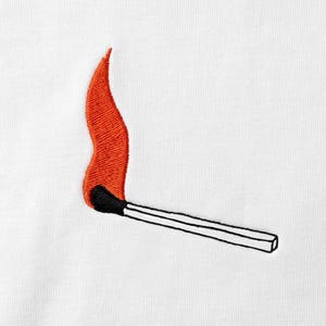 Puede incluir: Una superficie de tela blanca con un diseño bordado de una cerilla. La cerilla tiene una cabeza negra y una llama de color naranja vibrante que se extiende hacia arriba, con el palito de madera delineado en hilo negro sobre el fondo.