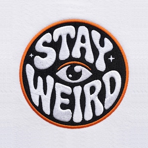 May include: A circular embroidered patch on a white fabric background. The patch features a black background with an orange border, white wavy lettering that reads STAY WEIRD, a central white eye graphic, and two small white four-pointed stars.