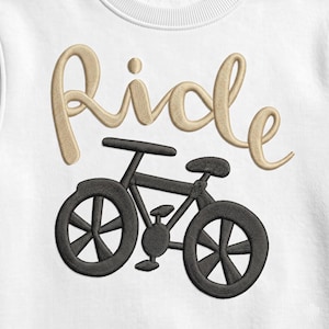 Può includere: Un capo bianco con un ricamo che raffigura una bicicletta nera posizionata sotto la parola ride, cucita in un carattere corsivo color marrone chiaro. Il ricamo ha un aspetto strutturato con dettagli del filo visibili nel disegno.