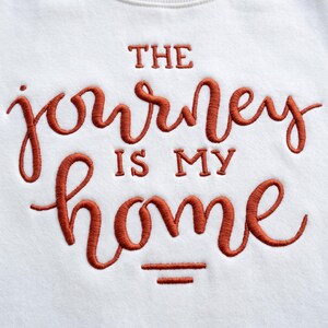 Może przedstawiać: Biała bluza z haftem w kolorze rdzy. Napis brzmi THE journey IS MY home, wykonany połączeniem liter drukowanych i kursywy, z dwiema poziomymi liniami umieszczonymi bezpośrednio pod słowem home.