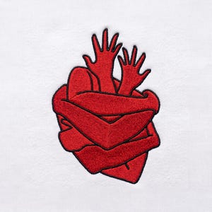 May include: A white fabric garment featuring a red embroidered patch. The design depicts two red arms wrapping around a central red shape, with two red hands extending upward from the top, all outlined in thin black thread.