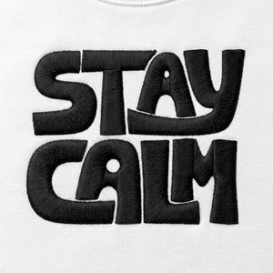 May include: A white fabric garment featuring the text STAY CALM embroidered in thick, black thread. The lettering is bold, rounded, and sans-serif, with a visible stitched texture throughout the black characters against the plain white background.