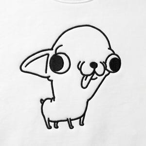 Puede incluir: Una sudadera blanca con un diseño bordado en hilo negro que representa a un perro pequeño con ojos circulares grandes, una oreja apuntando hacia un lado y la lengua fuera. El bordado consiste en contornos negros gruesos sobre tela blanca.