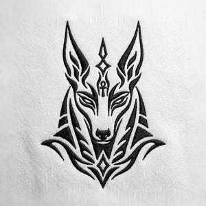 May include: A black embroidered design on a white fabric surface featuring the stylized head of an Egyptian jackal deity. The artwork includes sharp, angular lines, pointed ears, and an ankh symbol centered on the forehead above the nose.