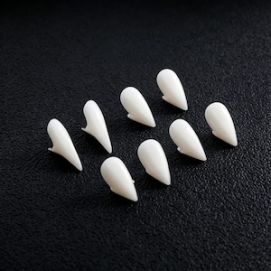 May include: A set of eight individual white plastic vampire fangs arranged in a scattered pattern. Each piece features a smooth, curved, pointed shape designed to resemble a canine tooth, with a small base tab for placement against a tooth surface.
